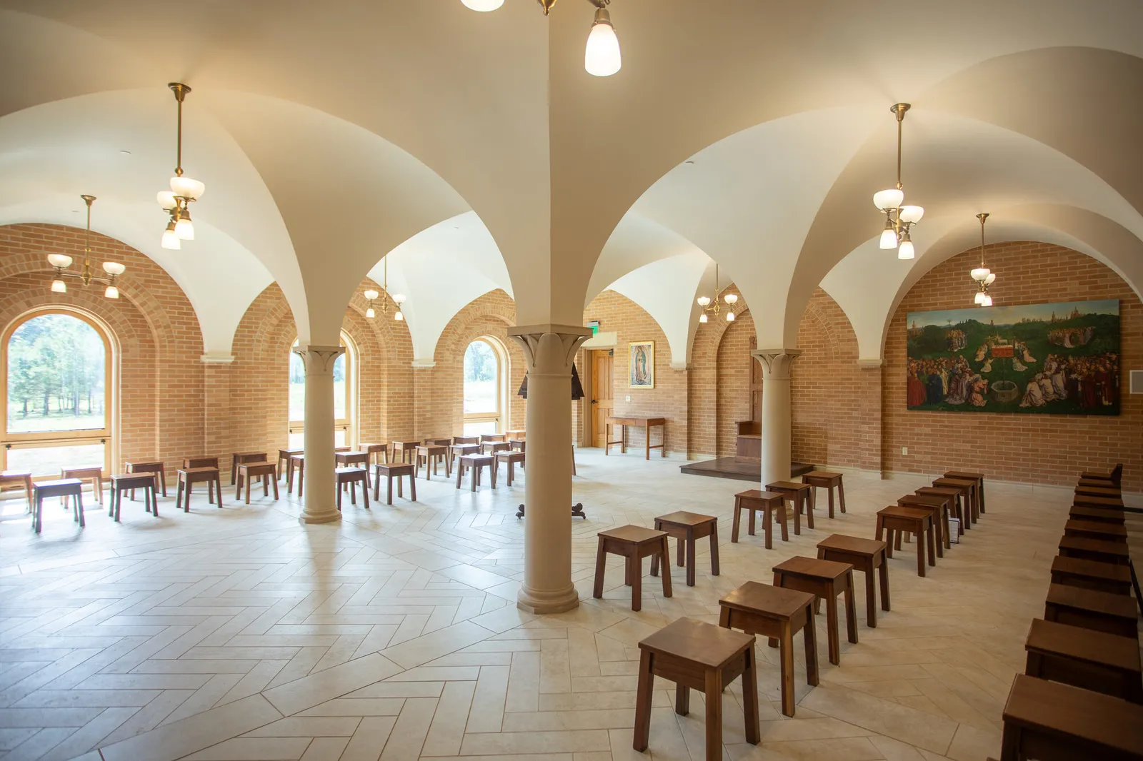 Photograph of a vaulted stone-columned hall with a herringbone-patterned floor, arched brick niches, and wooden stools arranged for gathering.