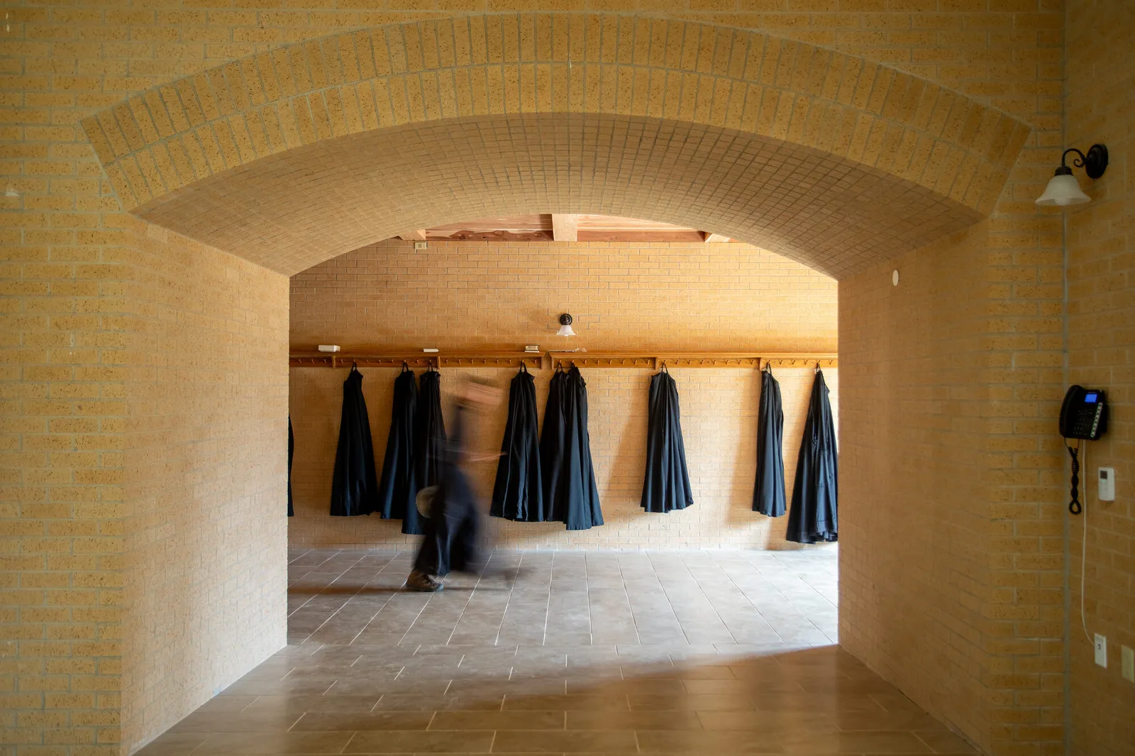 Photograph of a vaulted brick passage lined with hanging black choir robes, with a monk walking through in motion blur.