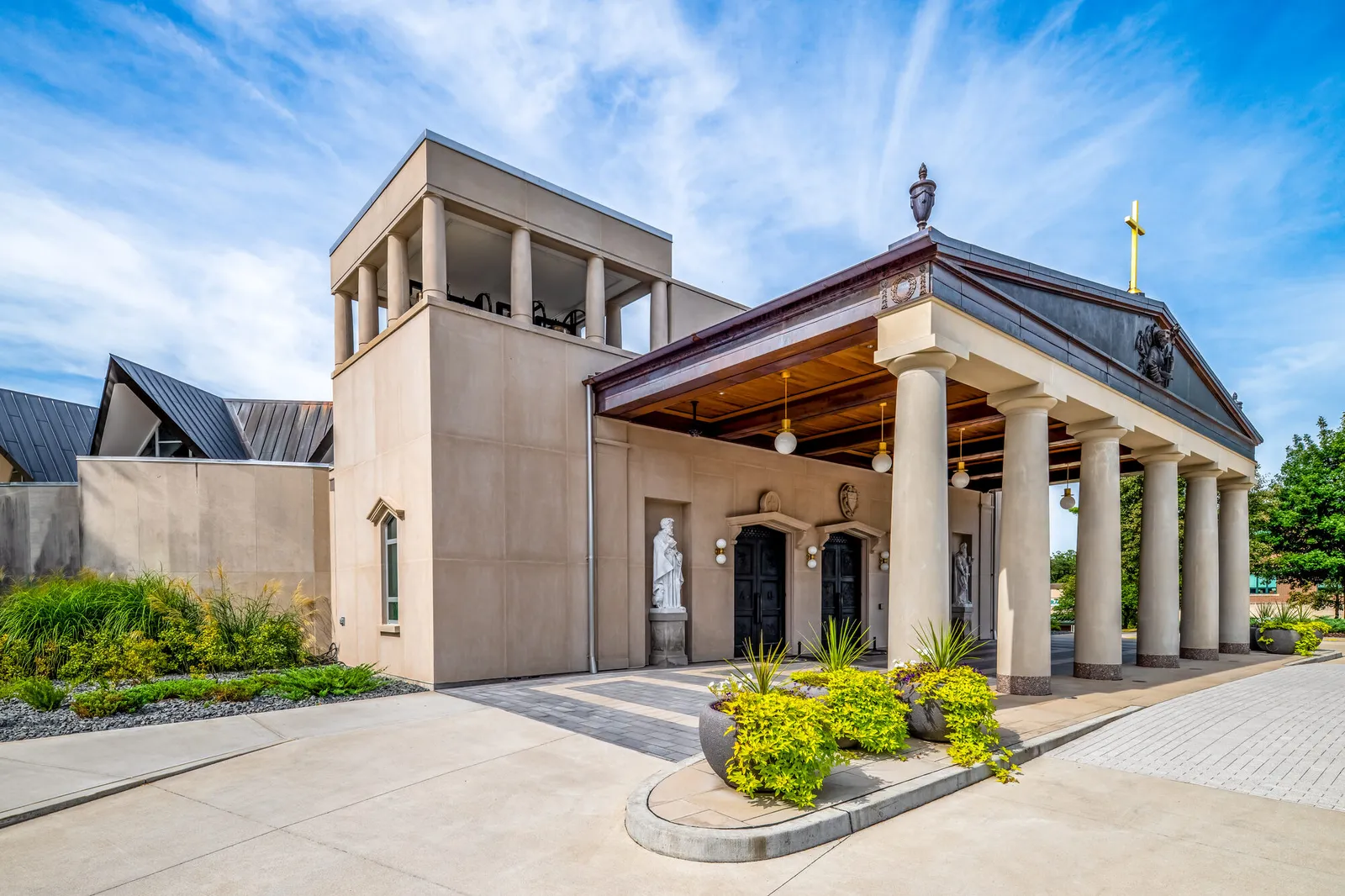 Photograph of the cathedral's stone entrance tower and Doric-columned portico, taken at an angle from the paved forecourt, with statues framed beneath the colonnade.