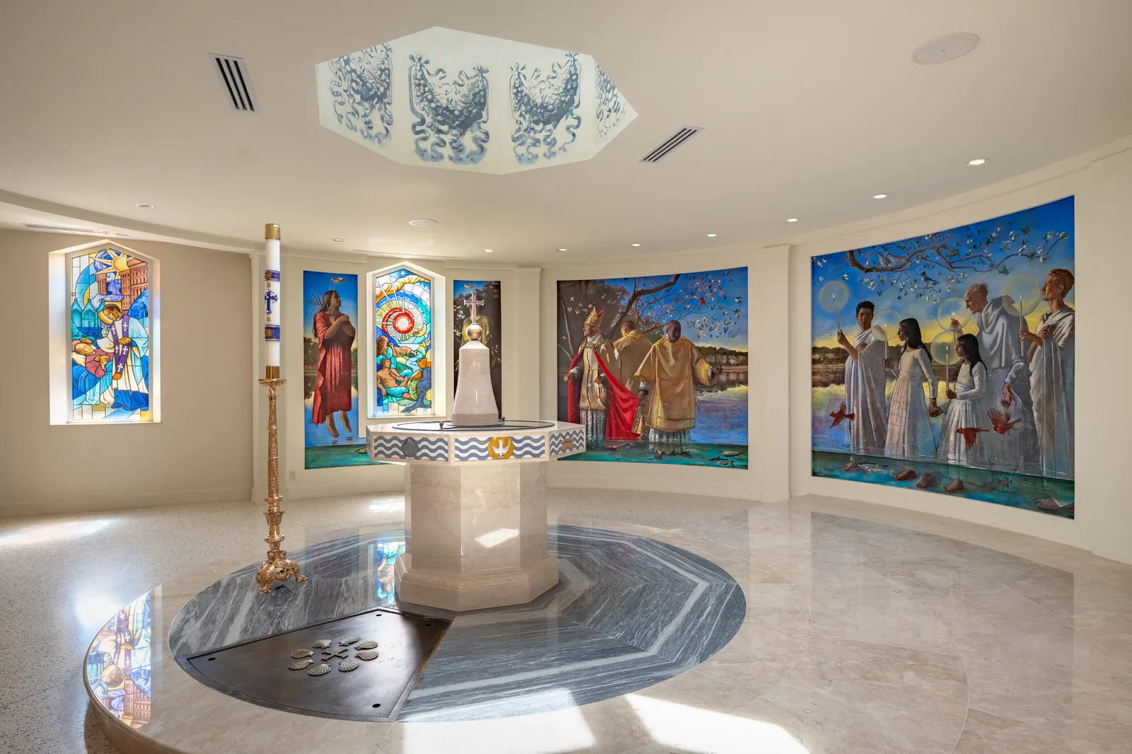 Interior photograph of a circular baptistery featuring an octagonal marble font, stained glass windows, painted murals of baptismal scenes, and a skylight with painted medallions.