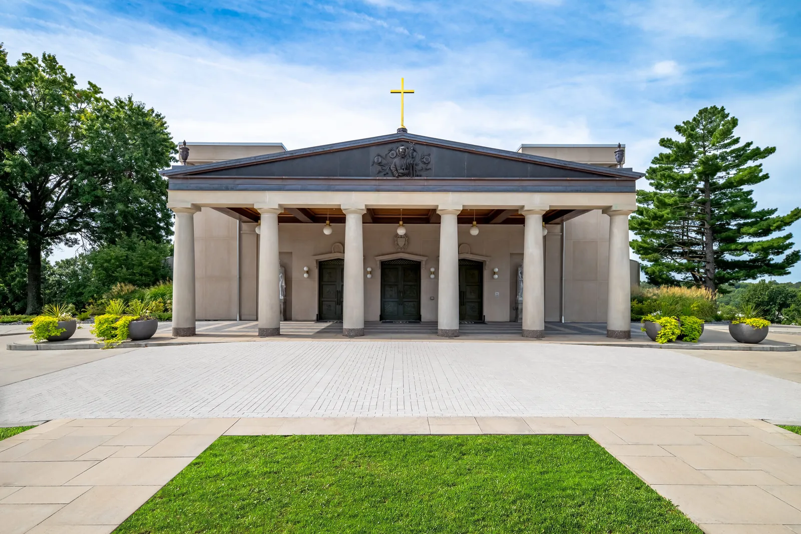 Symmetrical front elevation of the cathedral showing a six-column pedimented portico with a relief sculpture in the tympanum and a gilded cross above, framed by trees and a lawn.