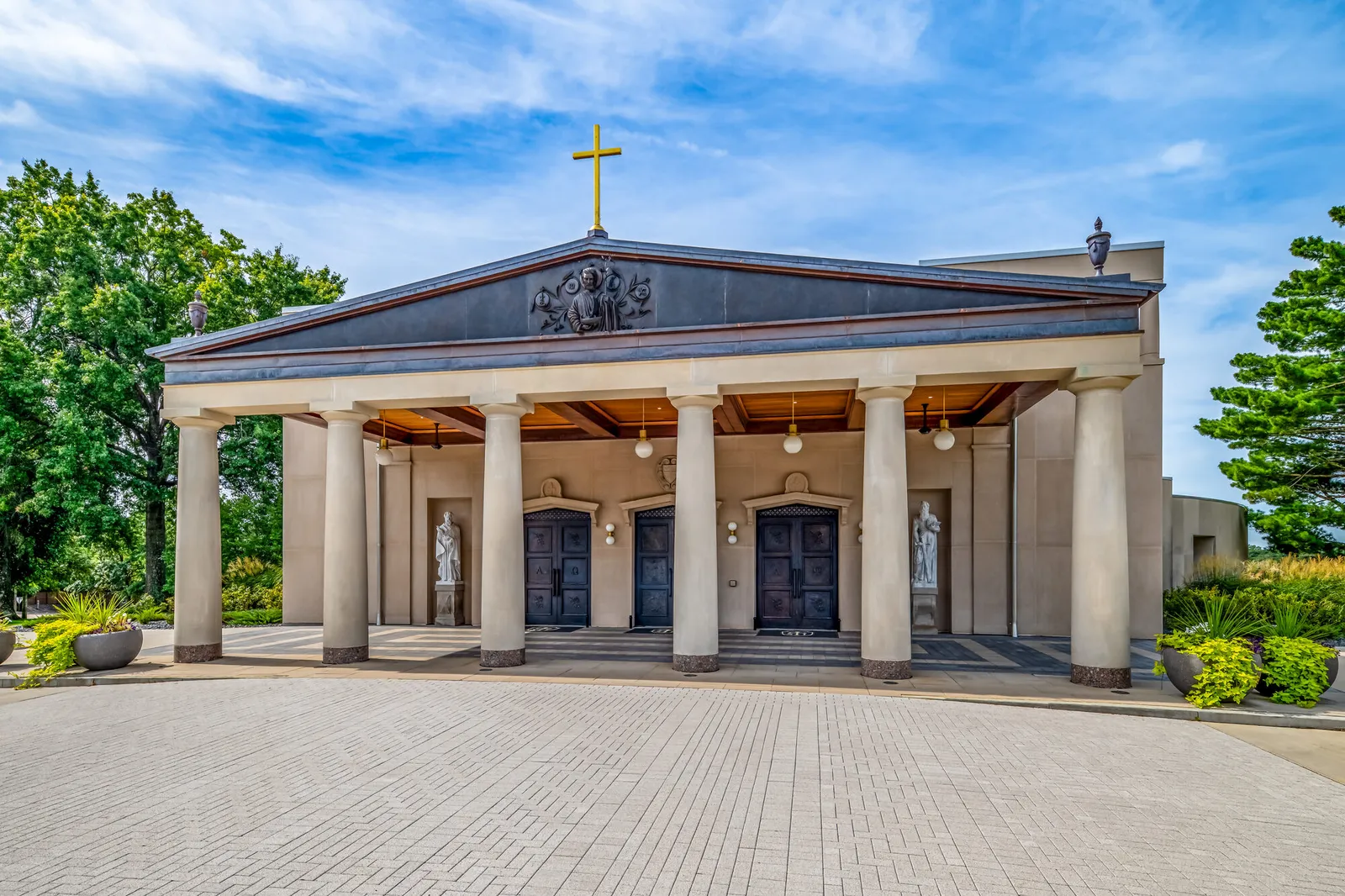 Front elevation of the cathedral's colonnaded portico with bronze doors, flanking statues, and a gilded cross atop the pediment, in warm afternoon light.