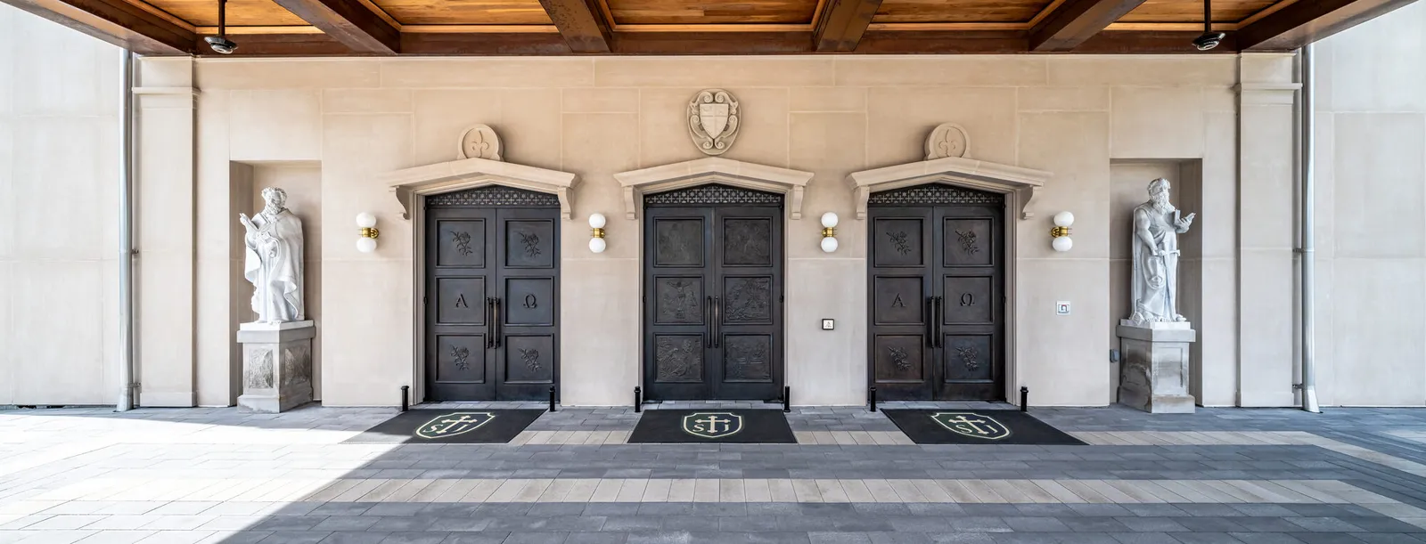 Close-up photograph of three sets of bronze entrance doors decorated with relief panels and alpha-omega motifs, flanked by stone statues on pedestals beneath a coffered wood ceiling.
