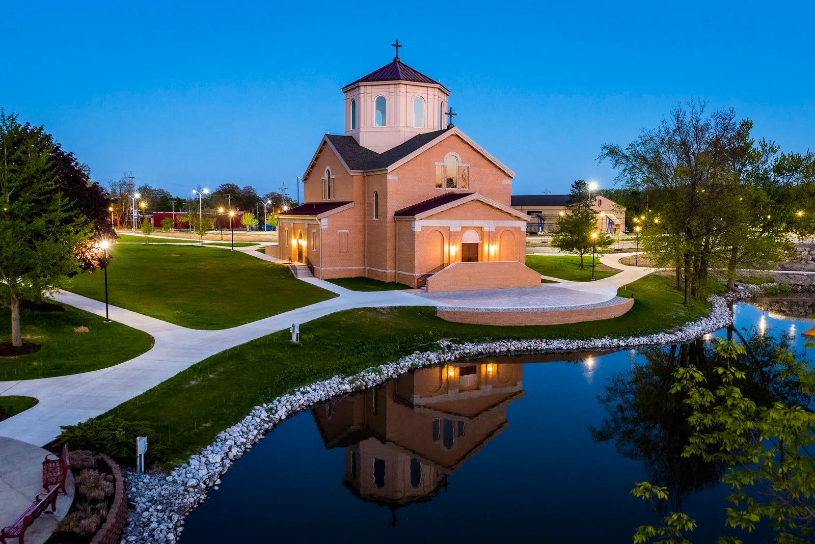 Twilight exterior view of a brick chapel with an octagonal lantern tower, illuminated within, reflected in a still pond surrounded by campus paths and lamps.
