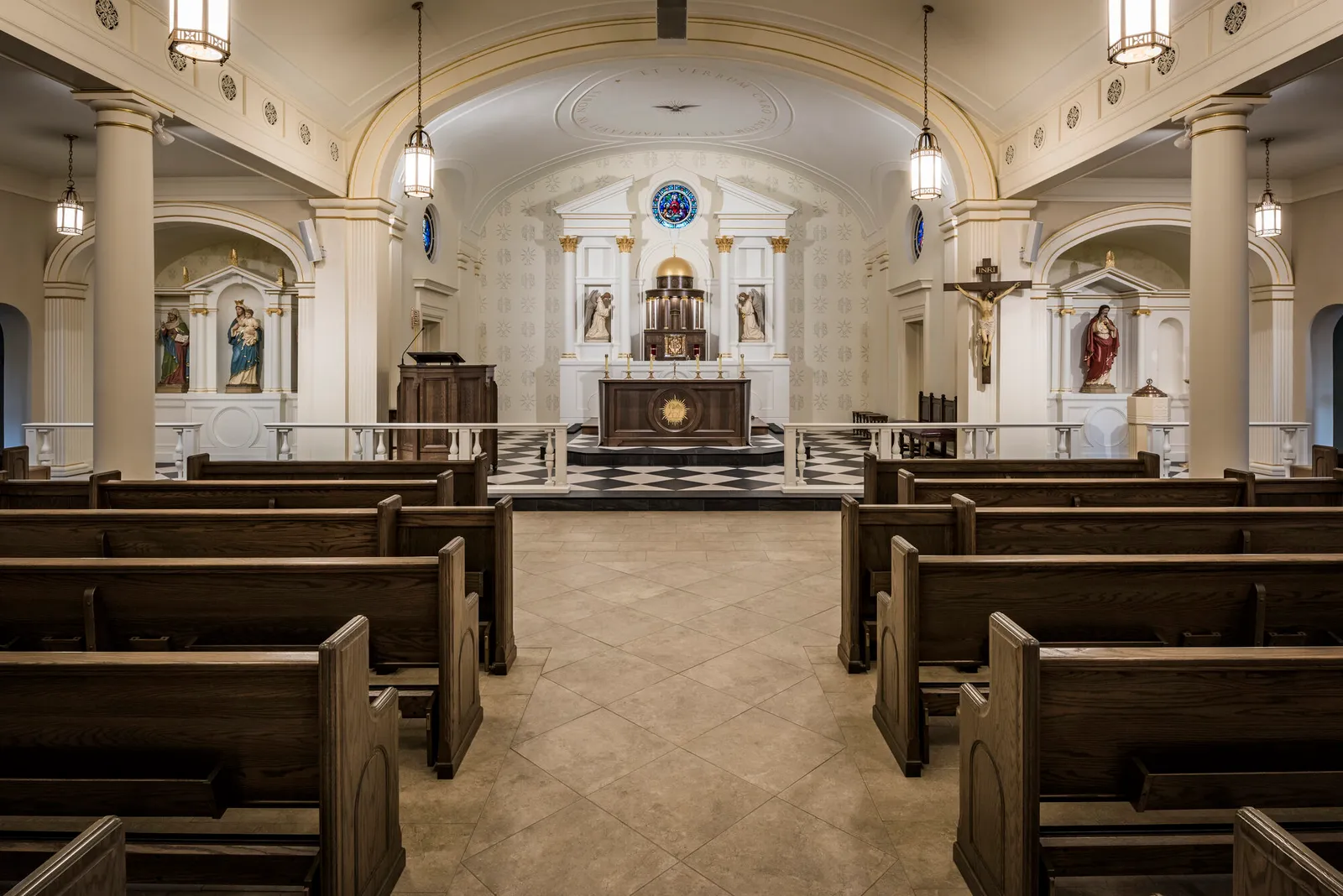 Interior of Blessed Sacrament looking down the nave toward the altar, with pendant lanterns, statue niches, and checkerboard flooring at the sanctuary.