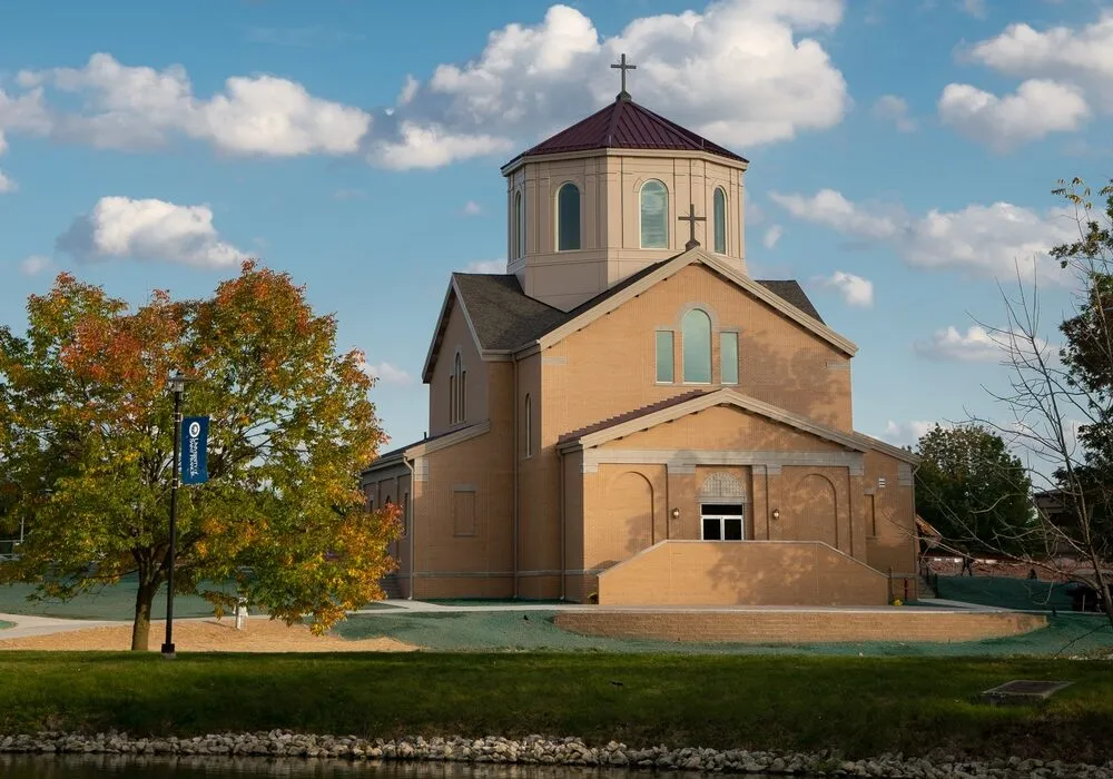 Daytime exterior view of a brick chapel with an octagonal lantern tower, with an autumn tree and a university banner on a lamp post in the foreground.