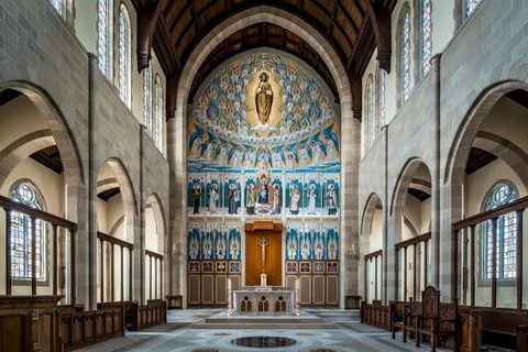 Interior photograph of a stone Gothic-style chapel nave with ribbed vaulting, stained glass windows, and a richly painted and mosaicked sanctuary wall depicting Christ enthroned among angels and saints.