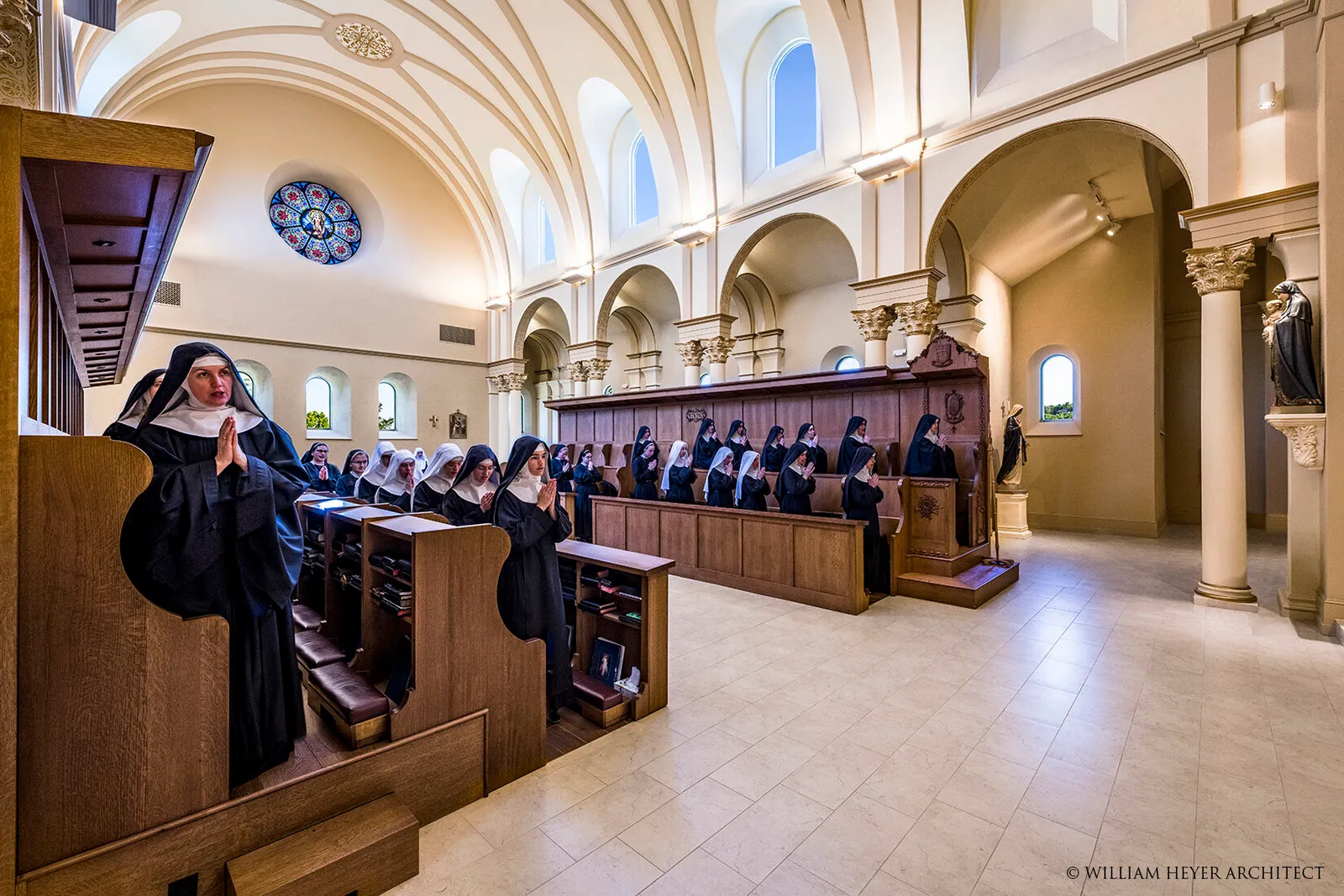 Benedictine nuns standing in wooden choir stalls with hands joined in prayer inside a vaulted chapel with high windows.
