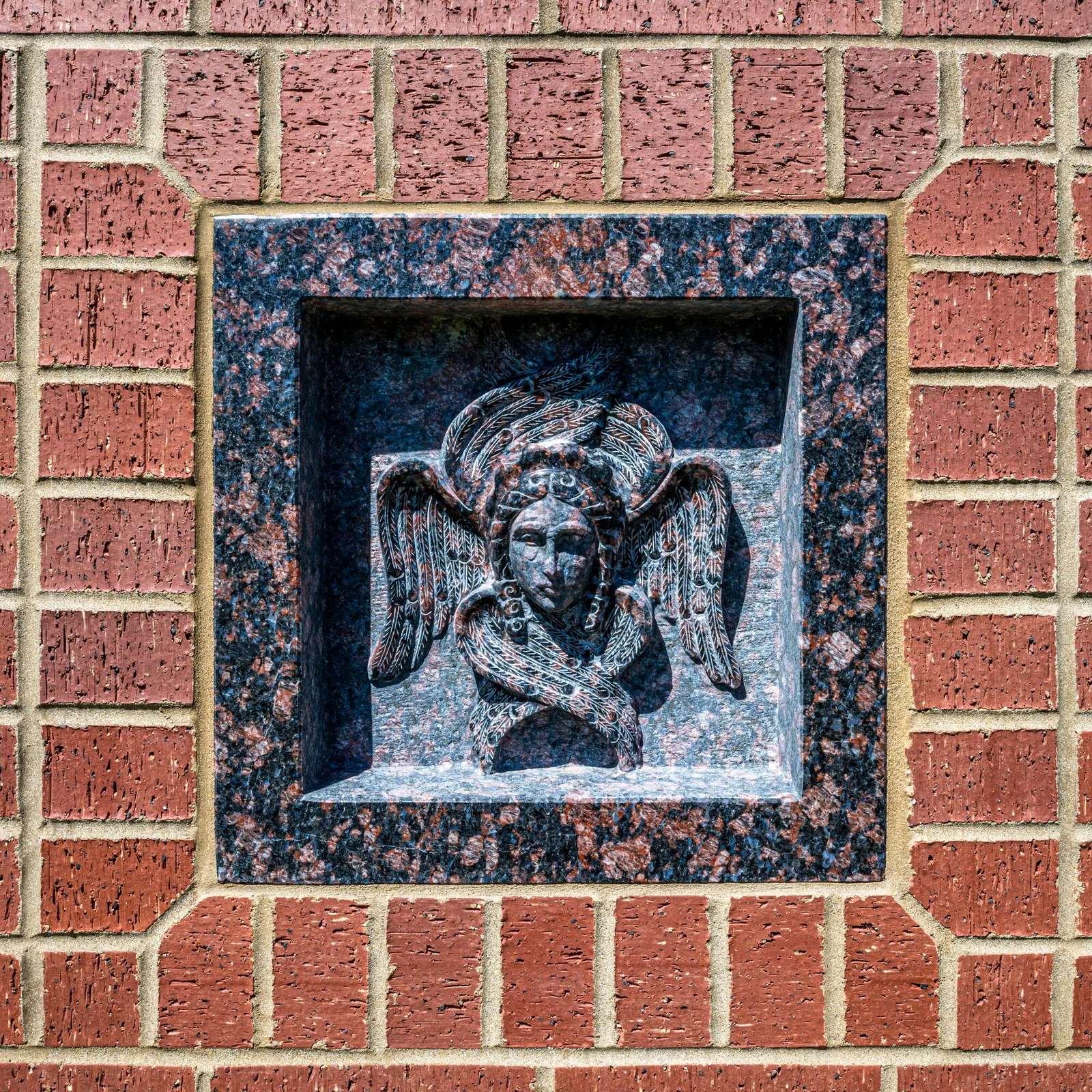 Close-up of a carved stone angel bust relief framed in polished granite within a brick wall.