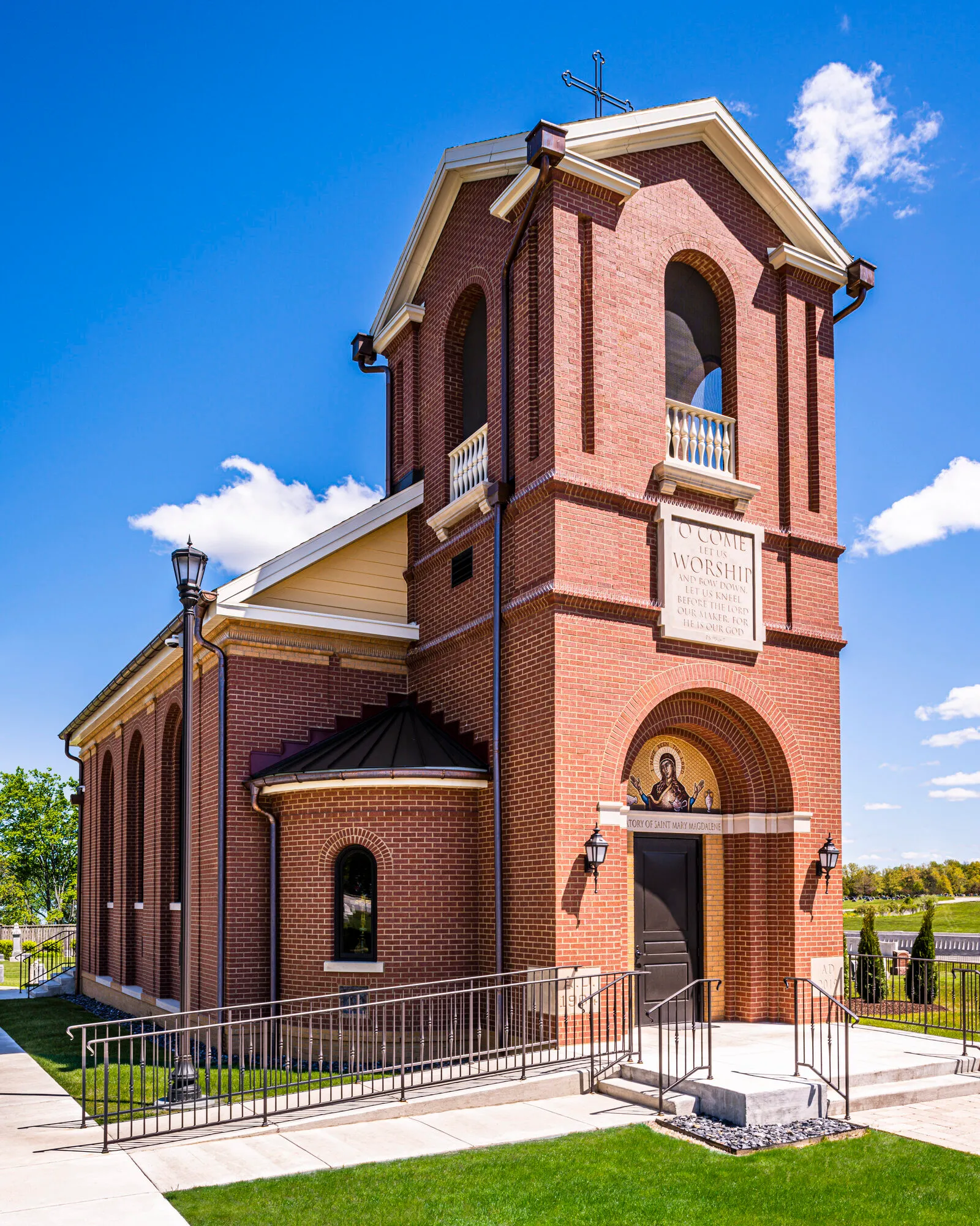 Angled exterior view of a red brick chapel bell tower with an arched belfry opening, inscribed stone plaque, and arched entrance below.