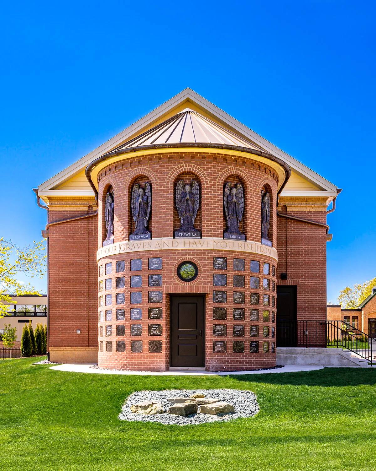 Symmetrical frontal view of a round brick columbarium building with five carved angel reliefs above rows of inscribed memorial plaques and a stone garden in front.