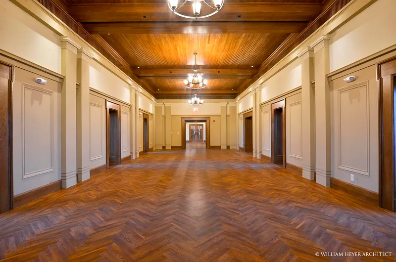 The same space completed: a herringbone wood floor between panelled walls with fluted pilasters, a coffered wood ceiling and brass chandeliers, doors to the faculty offices either side.