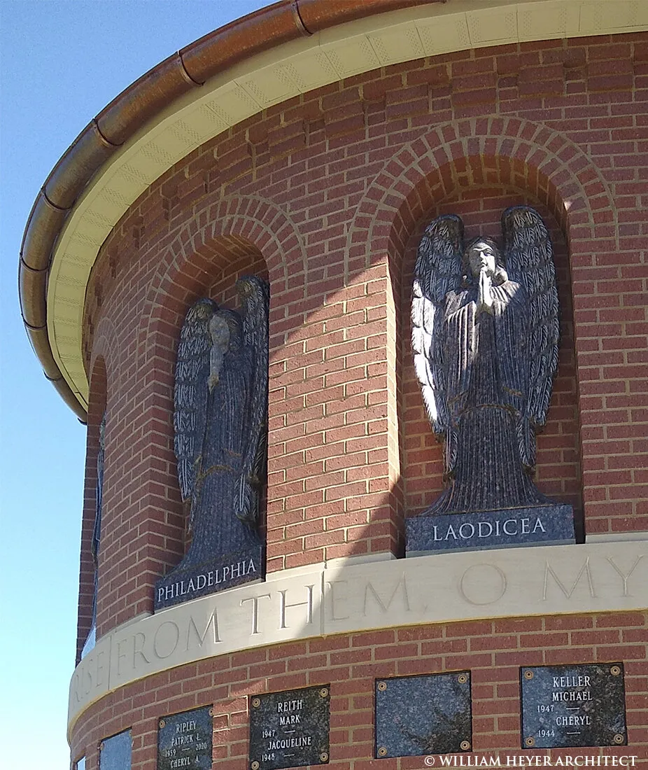 Two carved angels in the arched niches of the brick columbarium apse, above rows of memorial plaques and an inscribed stone band.