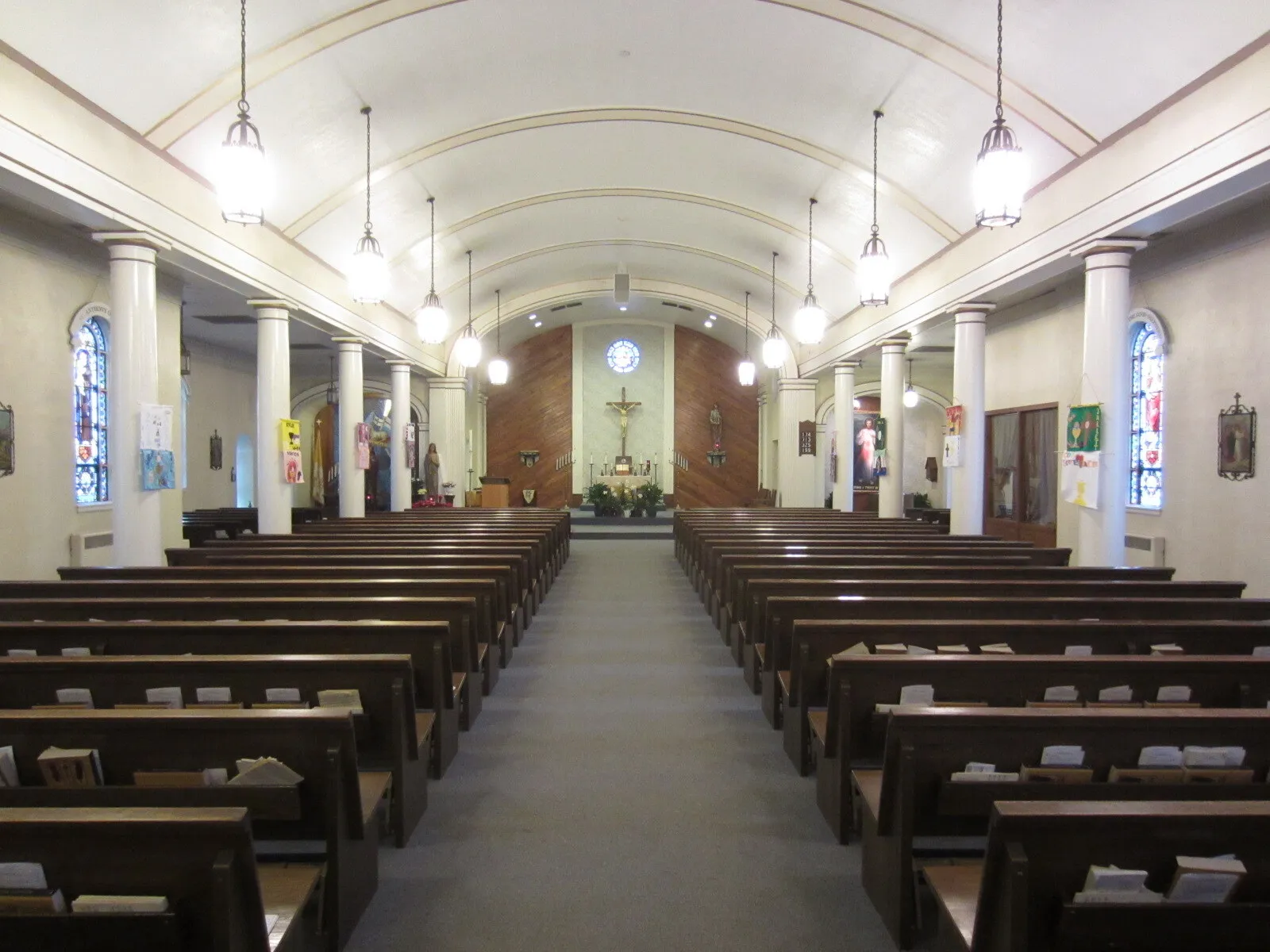 The nave before renovation, seen down the center aisle: a plain white barrel-vaulted ceiling, white columns, hanging lanterns, gray carpet and wood pews leading to a wood-paneled sanctuary.