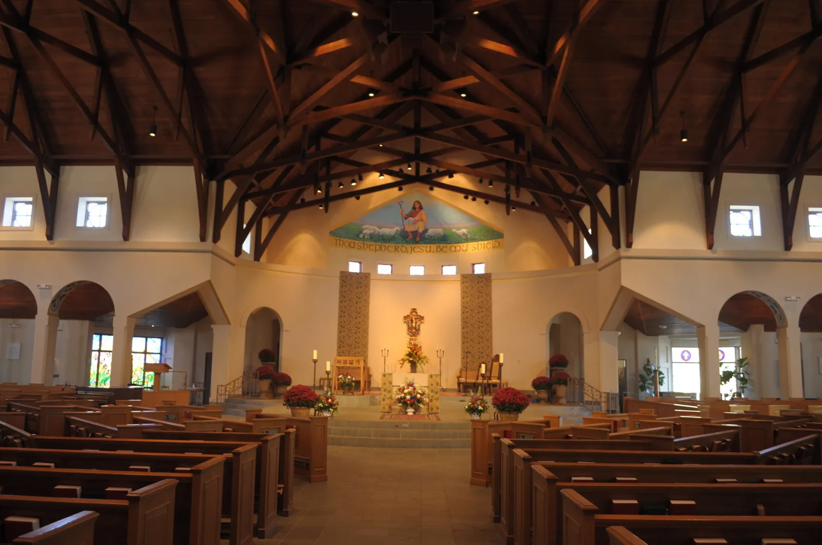 The existing church interior from the center aisle: exposed timber roof trusses, white walls, a mosaic of the Good Shepherd above the sanctuary, and wood pews.