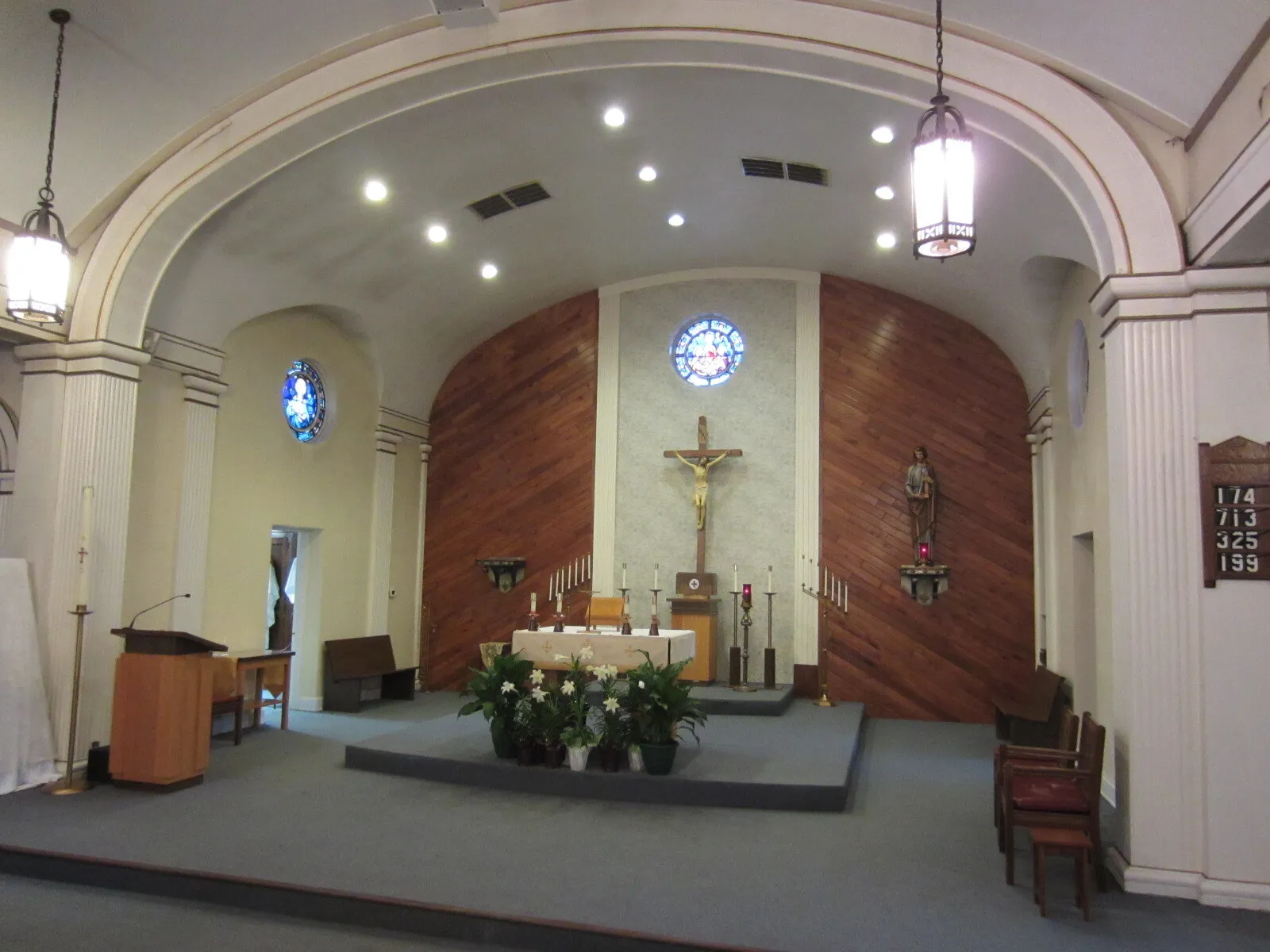 The sanctuary before renovation, with diagonal wood-plank paneling behind a simple altar, a crucifix, a small round stained-glass window, carpeted steps and recessed ceiling lights.