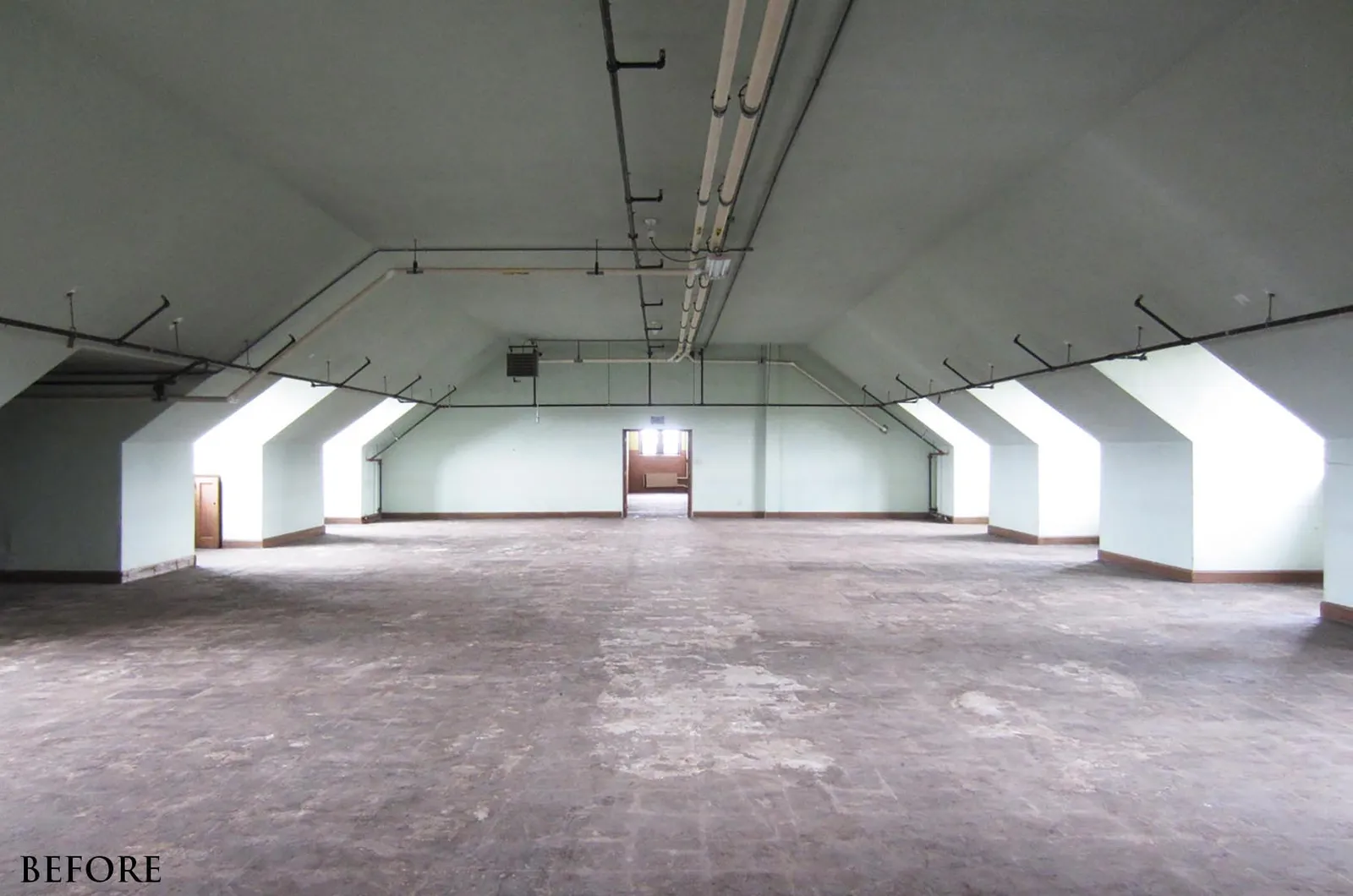 The attic space as found: a bare concrete floor under a low sloping ceiling with exposed ducts and pipes, dormer windows along both sides and a single door at the far end.