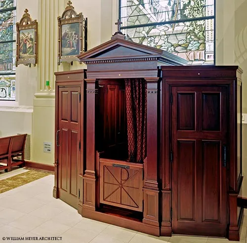 Mahogany confessional with pediment and paneled doors beside stained-glass windows