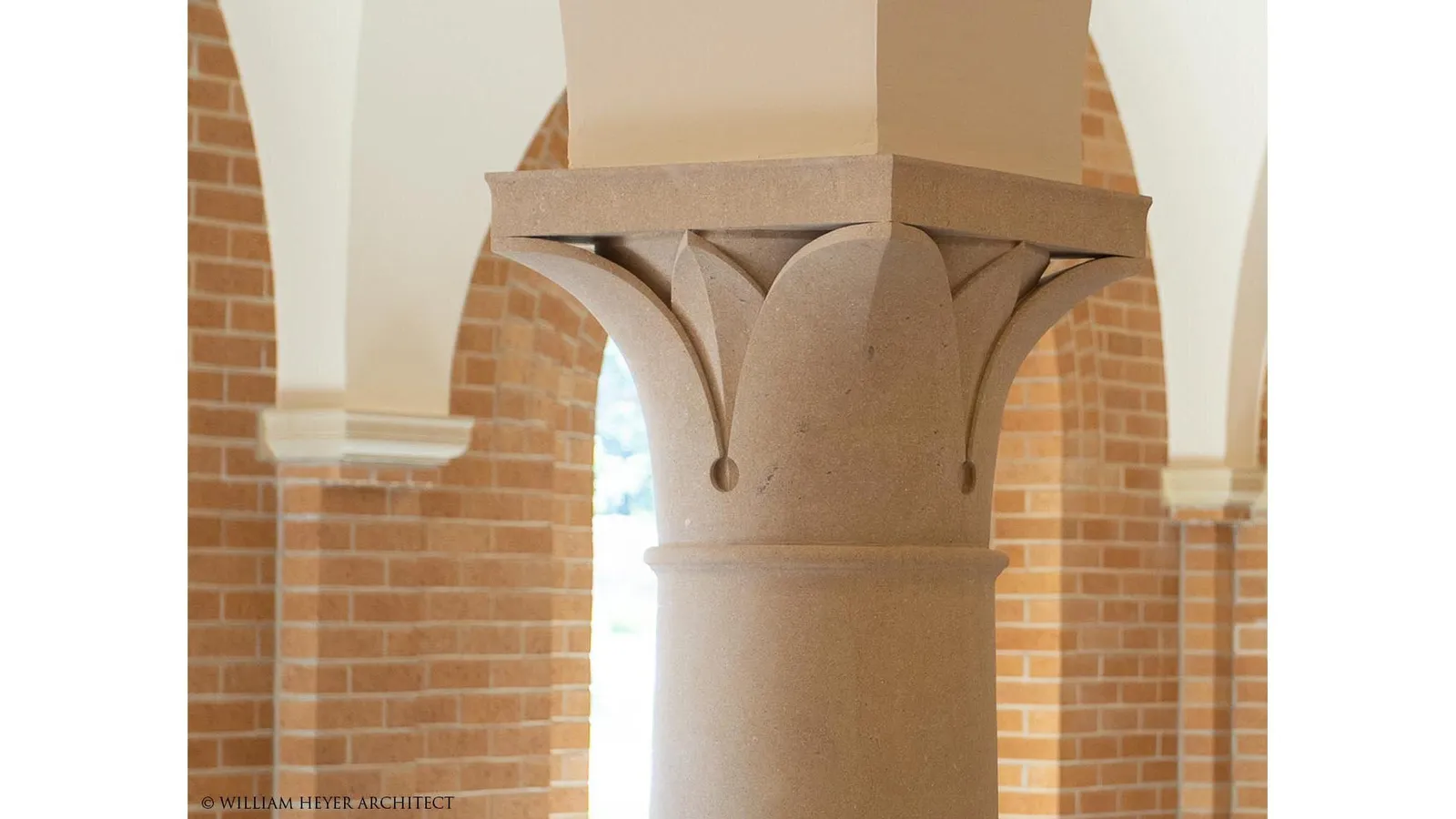 Close view of a stone column capital in the chapter room, a cushion form carved with a pointed leaf beneath a plain abacus and brick arches.