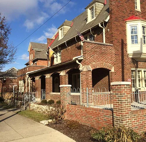 Brick clubhouse with arched porch, dormers, and flags along a residential sidewalk