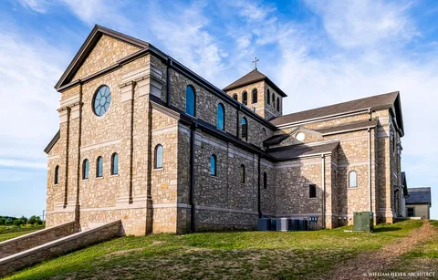 Exterior corner view of the stone abbey church showing the gable end with a rose window and the bell tower topped with a cross.