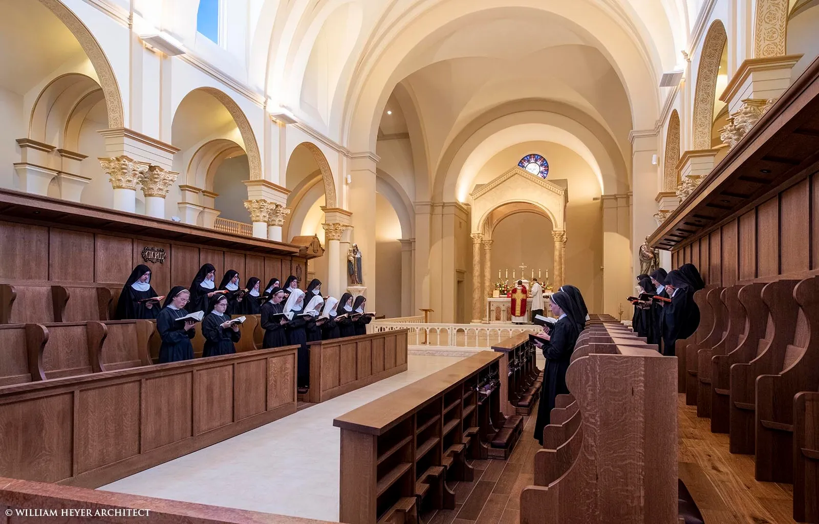Benedictine sisters singing from choir books in wooden stalls, with a priest at the altar visible in the background.