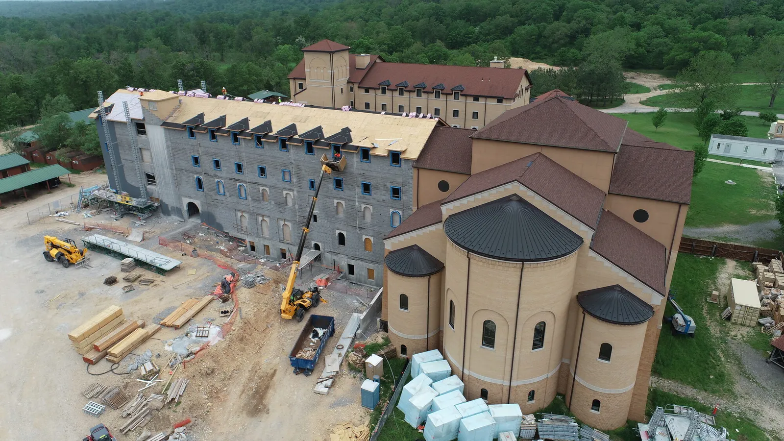 Aerial photograph of a monastery building under construction, with exposed concrete block walls, roof framing, and construction equipment, adjacent to the completed abbey church.