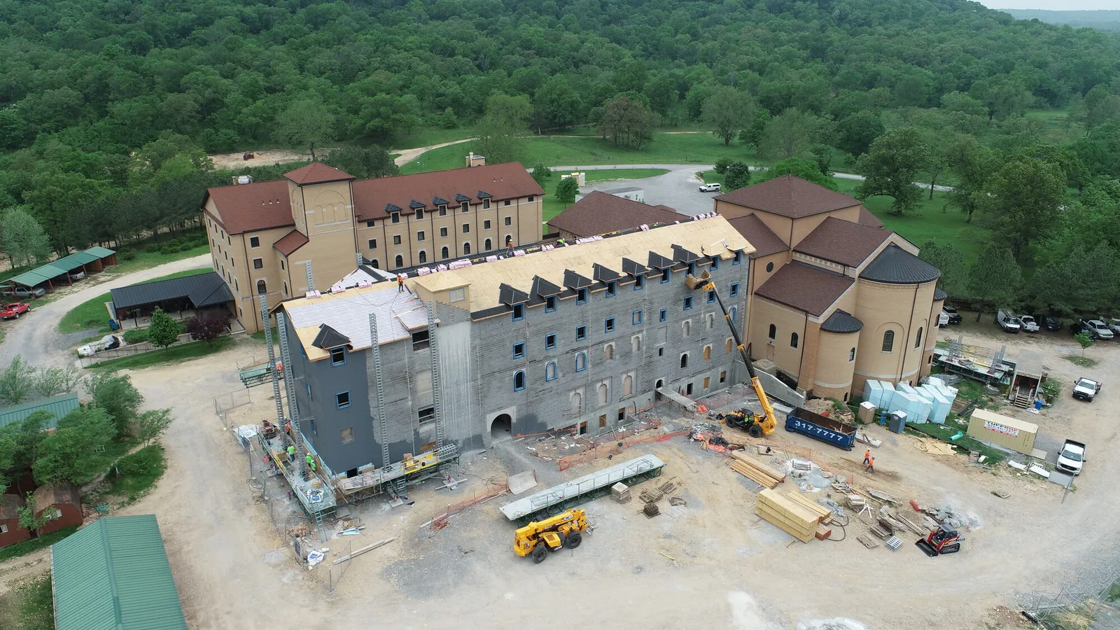 Aerial photograph of a monastery complex showing a partially completed dormitory wing under construction beside the finished abbey church and older buildings, surrounded by wooded hills.
