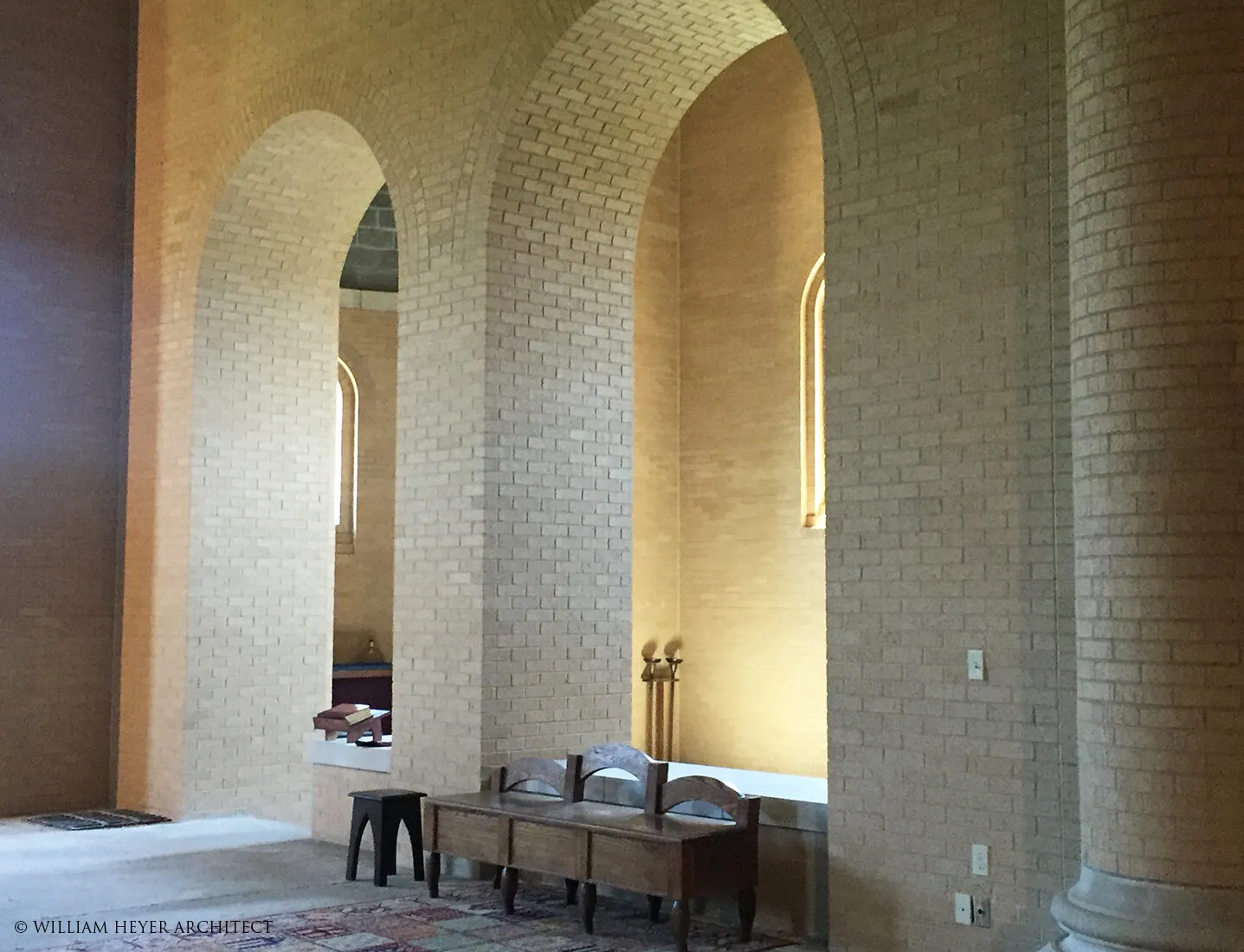 Pale brick piers and round arches inside the abbey, a bench and tall candlesticks set against the wall in raking light.