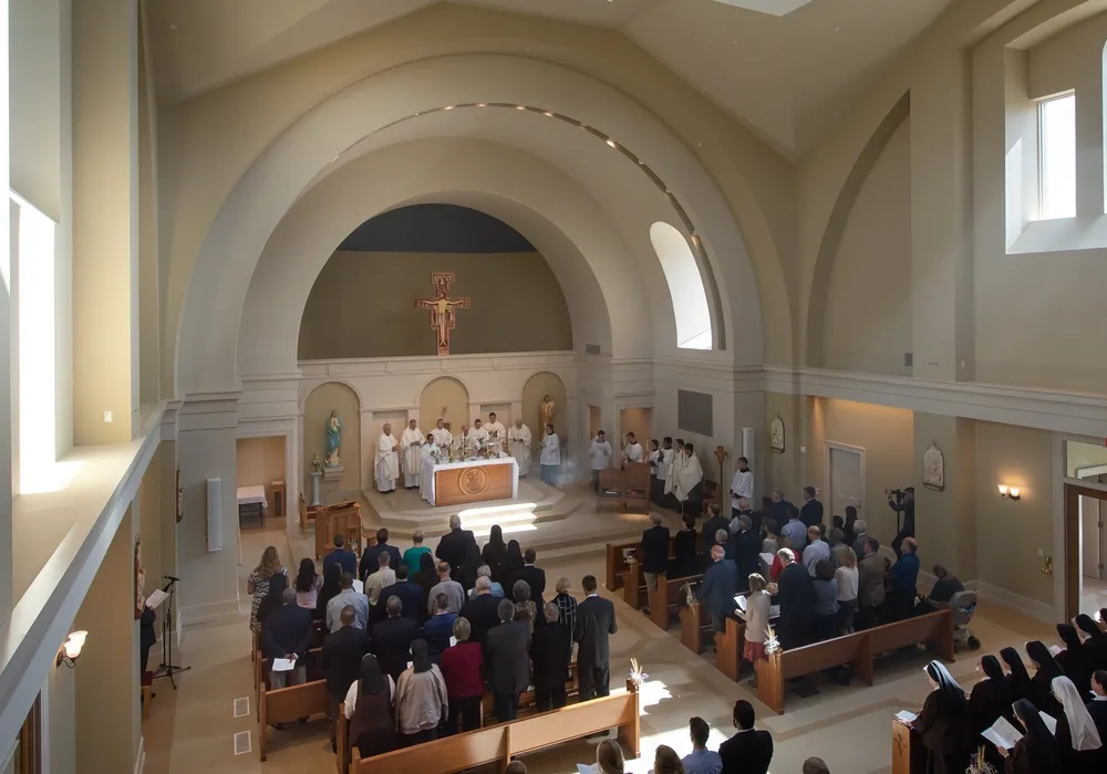 The completed oratory interior during Mass, seen from the gallery: a barrel-vaulted nave with plain cream walls, a rounded apse framed by pilasters with a painted crucifix above the altar, clergy at the altar and a full congregation standing in wooden pews.