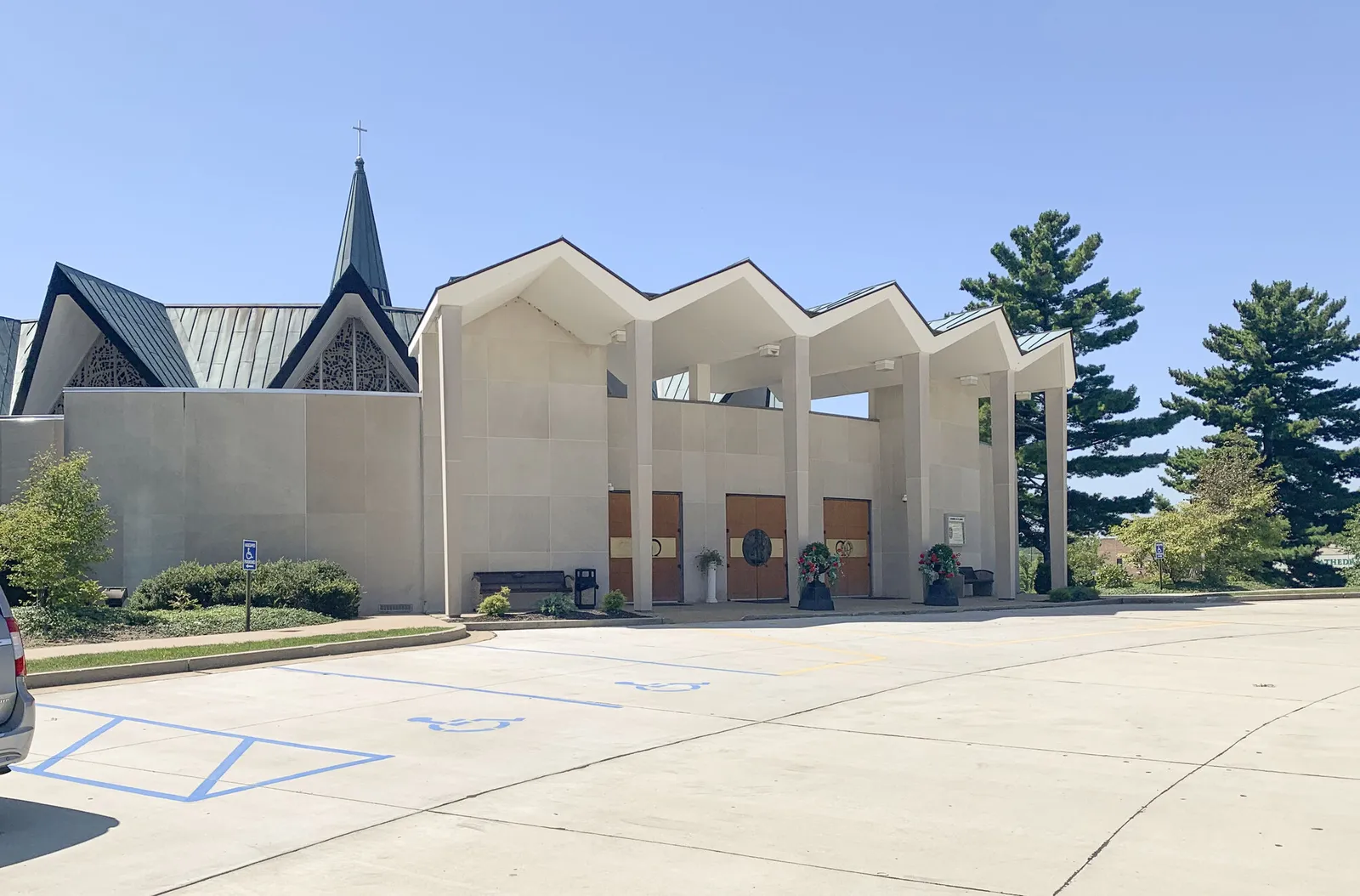 The cathedral entrance before renovation: a zigzag-roofed canopy on slender columns over three wood doors, with the green metal-roofed church and spire behind, seen across the parking lot.