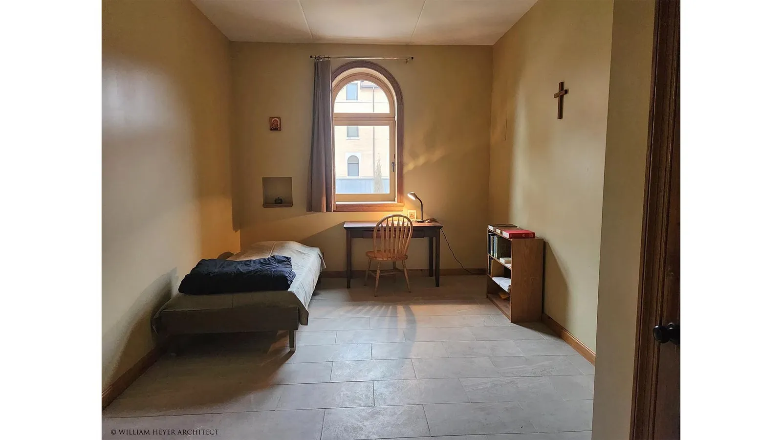A monk's cell: a bed, a desk and chair beneath an arched window, a crucifix on the wall and a stone floor.