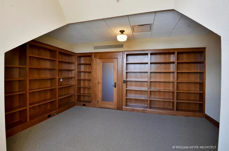A faculty office fitted with floor-to-ceiling wooden bookshelves across the end wall around a panelled door, under the sloping attic ceiling.