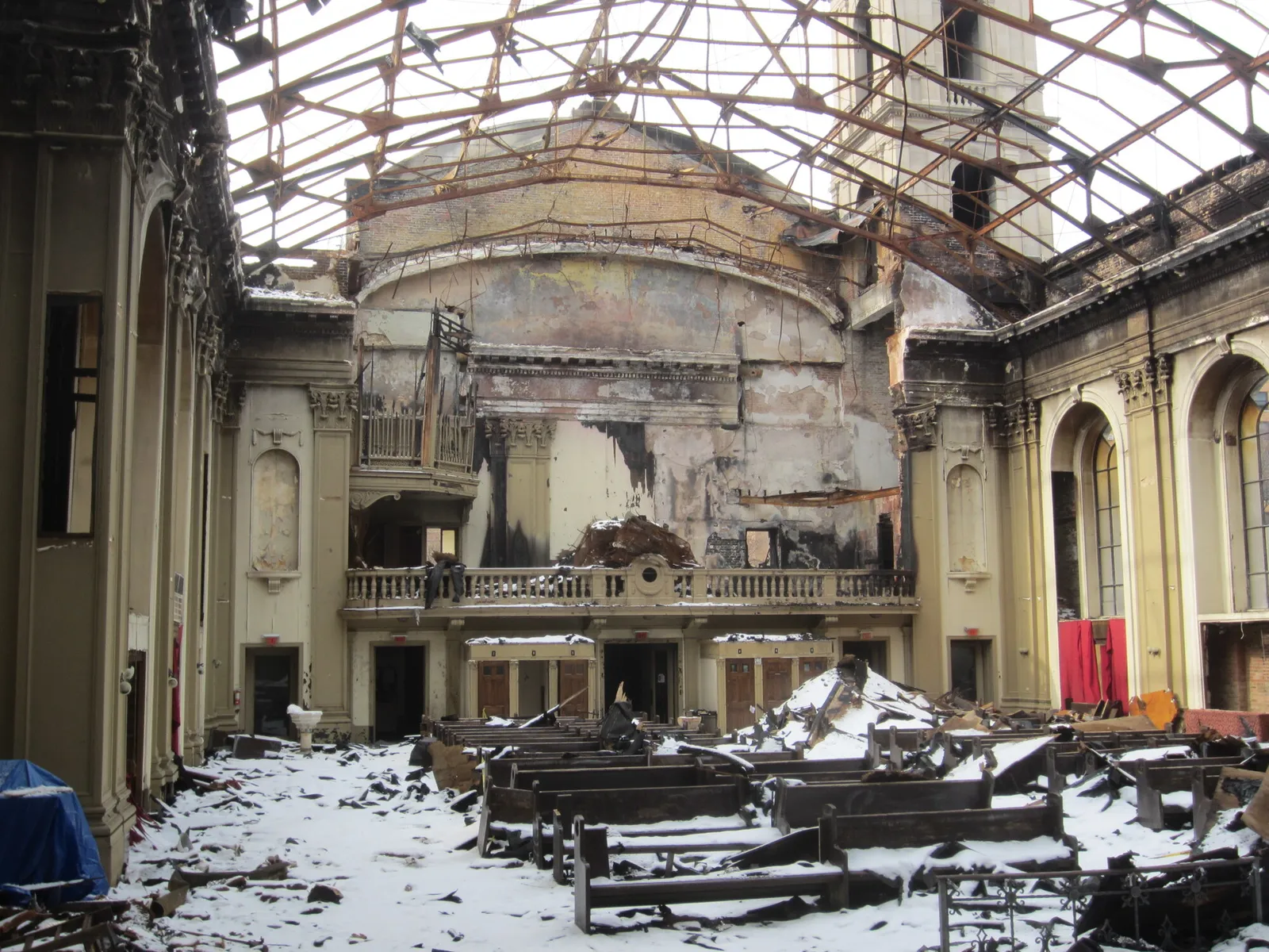 The fire-gutted nave toward the choir loft, open to the sky through the rusted roof frame, with charred walls and snow lying on the debris and pews.