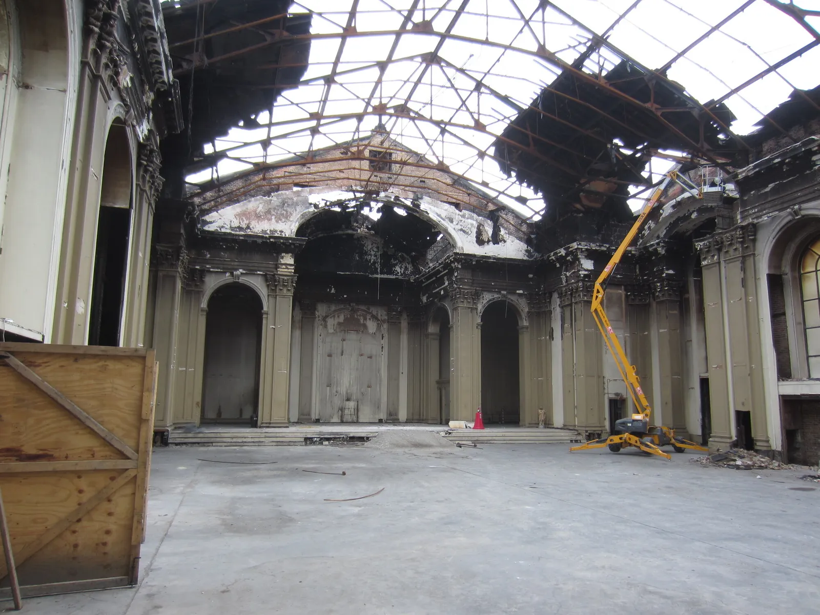 The fire-gutted sanctuary end of the church, open to the sky through the burnt roof frame, with scorched plaster, a bare concrete floor and an articulating lift.