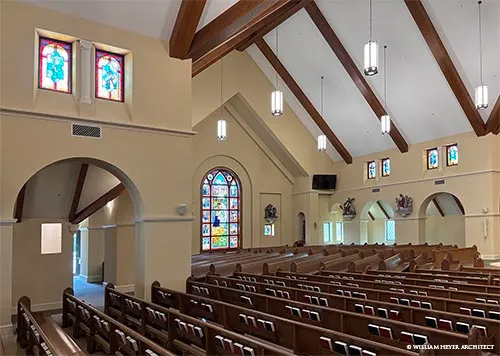 Church interior with exposed timber trusses, pendant lights, and stained-glass windows over pews