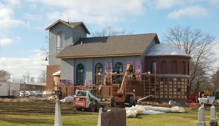A brick chapel with bell tower and apse under construction, scaffolding along its front with a lift and skid loader in the foreground